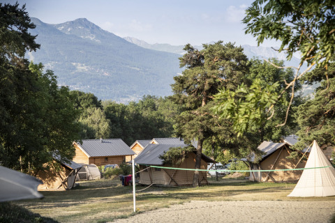Hütten und Zelte unter Bäumen mit Blick auf Berge im Huttopia Bourg Saint-Maurice, Auvergne-Rhône-Alpes, Frankreich.