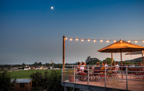 Outdoor dining area with guests under string lights and parasol at Huttopia Meursault, Bourgogne, France.