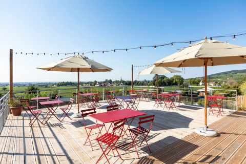Terraza al aire libre con mesas y sillas rojas, sombrillas y vistas panorámicas en Huttopia Meursault, Francia.