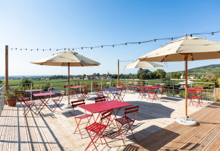 Buitenterras met rode tafels, stoelen, grote parasols en uitzicht op Huttopia Meursault, Bourgogne, Frankrijk.