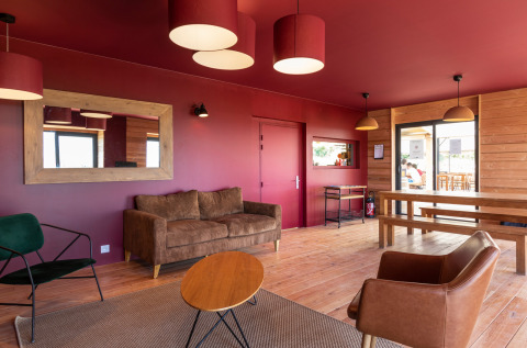 Salón moderno con paredes rojas, muebles de madera y grandes ventanales en Huttopia Meursault en Francia.
