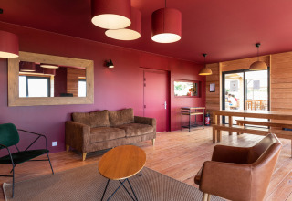 Modern lounge with red walls, wood furnishings, and large windows at Huttopia Meursault holiday park in France.