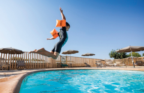 Niño con flotadores salta a la piscina en Huttopia Meursault, parque vacacional bajo cielo soleado.