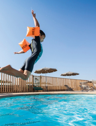 Niño con flotadores salta a la piscina en Huttopia Meursault, parque vacacional bajo cielo soleado.