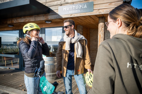 Tres personas sonríen frente a Huttopia Meursault en Bourgogne-Franche-Comté, Francia; una lleva casco de bicicleta.