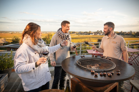 Tres personas disfrutan de vino y asan castañas al aire libre con vistas a viñedos en Huttopia Meursault.