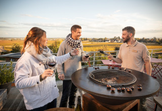 Tre persone gustano vino e arrostiscono castagne all’aperto con vista sui vigneti a Huttopia Meursault.