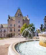 Fuente frente a un castillo histórico en Meursault, Bourgogne-Franche-Comté, Francia, bajo cielo azul.