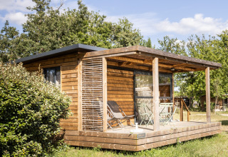 Træhytte med terrasse og havemøbler i solskin, omgivet af grønne træer i Huttopia Vallée de la Semois, Namur.