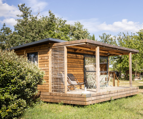 Wooden cabin with veranda and outdoor furniture in the sunlight, surrounded by trees at Huttopia Vallée de la Semois.