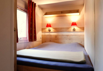 Kleines Schlafzimmer mit Doppelbett, roten Lampen, Fenster und Vorhängen in einer Hütte bei Huttopia Royat.