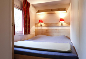 Kleines Schlafzimmer mit Doppelbett, roten Lampen, Fenster und Vorhängen in einer Hütte bei Huttopia Royat.