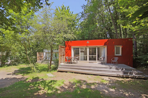 Cabane mobile rouge avec terrasse en bois au milieu des arbres à Huttopia Royat, en France.