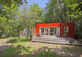 Rotes Mobilheim mit Holzterrasse inmitten von Bäumen im Huttopia Royat, Frankreich, im Sommer.
