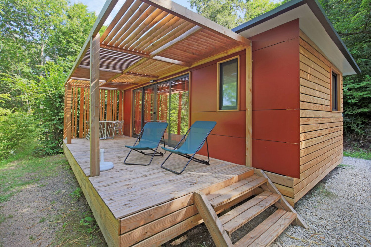 Casa móvil Vancouver en Huttopia Royat, Francia, con terraza de madera, sillas y rodeada de árboles.