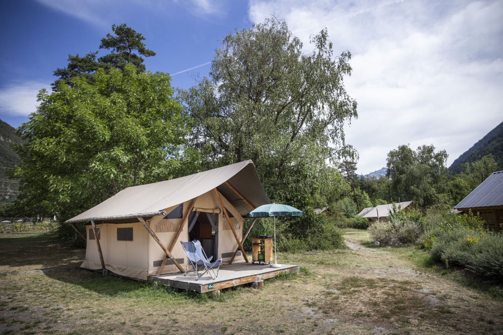 Safari Trappeur-tent met stoelen en parasol op houten terras, omringd door bomen en natuurgebied.