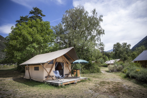 Tente safari Trappeur sur terrasse en bois avec chaises et parasol, entourée de verdure et d’arbres.