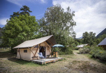Safari Trappeur-telt omgivet af træer og natur, med stole og parasol på en træveranda i campingområde.