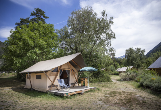 Safari Trappeur-telt omgivet af træer og natur, med stole og parasol på en træveranda i campingområde.