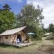 Safari Trappeur-telt omgivet af træer og natur, med stole og parasol på en træveranda i campingområde.