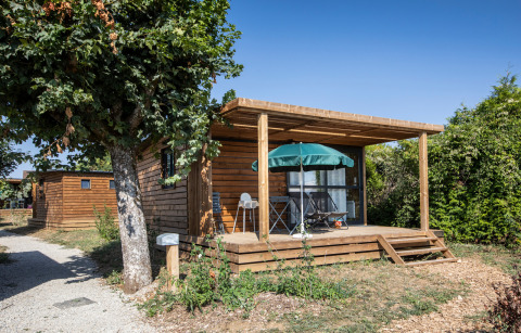 Cabaña Chalet Evasion de madera con terraza, mesa y sombrilla, rodeada de naturaleza y cielo azul.