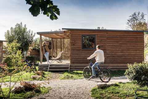 Man op fiets en vrouw bij Chalet Evasion te Huttopia Meursault, Frankrijk, op een zonnige dag.