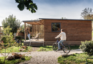 Hombre en bicicleta y mujer en la terraza del Chalet Evasion en Huttopia Meursault, Francia, al sol.