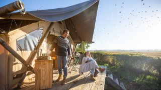Dos personas disfrutan café matutino en la terraza de una tienda safari Trapper II con paisaje soleado.