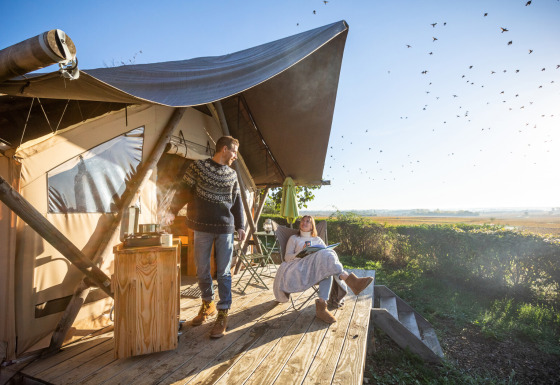 To personer nyder morgenkaffe foran en Trapper II safari-telt på en solrig terrasse med udsigt.
