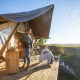 To personer nyder morgenkaffe foran en Trapper II safari-telt på en solrig terrasse med udsigt.