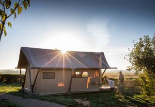 Trapper II safari tent at Huttopia Meursault in France, glowing in the sunrise with two people outside.