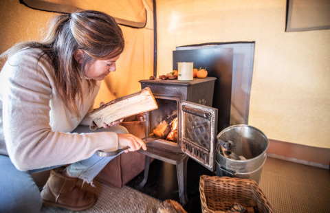 Mujer colocando leña en una estufa de leña en una tienda safari Trapper II en Huttopia Meursault, Francia.