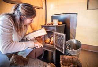 Mujer colocando leña en una estufa de leña en una tienda safari Trapper II en Huttopia Meursault, Francia.
