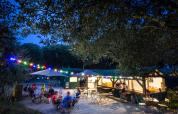 Udendørs café med folk, farverige lys og træer om aftenen på Huttopia Oléron Les Chênes Verts, Frankrig.
