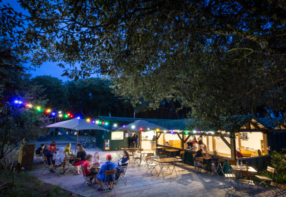 Udendørs café med folk, farverige lys og træer om aftenen på Huttopia Oléron Les Chênes Verts, Frankrig.