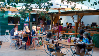 Familien essen gemeinsam draußen unter Sonnenschirmen im Ferienpark Huttopia Oléron Les Chênes Verts, Frankreich.