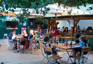 Familien essen gemeinsam draußen unter Sonnenschirmen im Ferienpark Huttopia Oléron Les Chênes Verts, Frankreich.