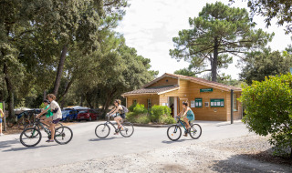 Huéspedes pasean en bicicleta frente a un edificio de madera en Huttopia Oléron Les Chênes Verts, Francia.