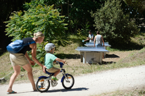 Una mujer ayuda a un niño en bicicleta mientras dos personas juegan al ping-pong al aire libre en Huttopia Oléron.