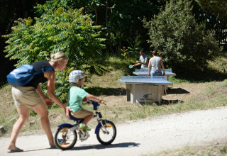En kvinde hjælper et barn på cykel, mens to personer spiller bordtennis i naturen ved Huttopia Oléron Les Chênes Verts.
