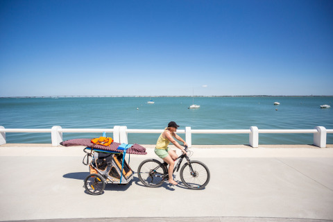 Frau fährt Fahrrad mit Anhänger voller Strandausrüstung am Meer bei Huttopia Oléron Les Chênes Verts.
