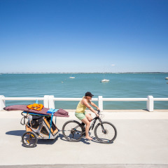 Frau fährt Fahrrad mit Anhänger voller Strandausrüstung am Meer bei Huttopia Oléron Les Chênes Verts.