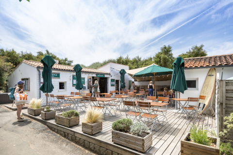 Terraza de cafetería al aire libre con mesas, sillas y sombrillas en Huttopia Côte Sauvage, Francia.