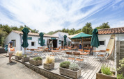 Udendørs caféterrasse med borde, stole og parasoller på Huttopia Côte Sauvage feriepark i Frankrig.