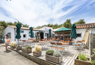 Terraza de cafetería al aire libre con mesas, sillas y sombrillas en Huttopia Côte Sauvage, Francia.