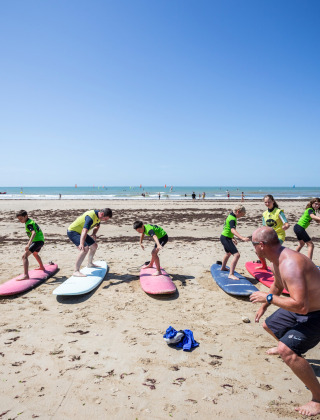 Children and adults take part in a surfing lesson on the beach near Sainte-Marie-de-Ré, France, under a clear sky.