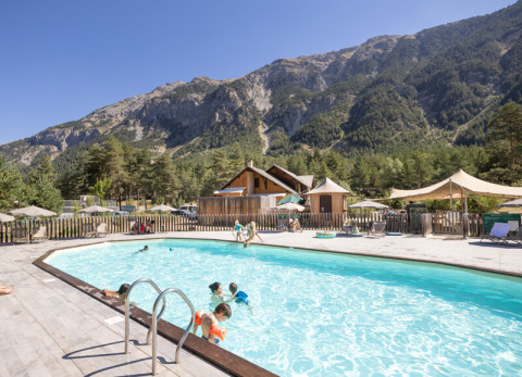 Piscina exterior en Huttopia la Clarée con niños jugando, rodeada de montañas y bosque en Francia.
