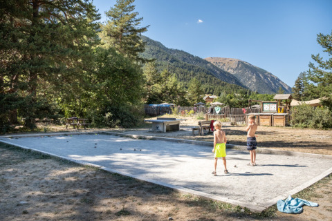Dos niños juegan petanca en una cancha de arena rodeada de árboles y montañas en Huttopia la Clarée, Francia.