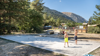 Dos niños juegan petanca en una cancha de arena rodeada de árboles y montañas en Huttopia la Clarée, Francia.