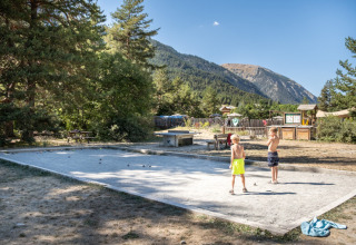 Dos niños juegan petanca en una cancha de arena rodeada de árboles y montañas en Huttopia la Clarée, Francia.
